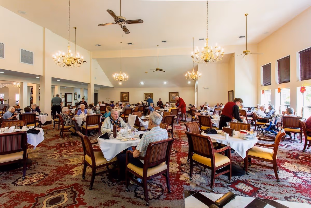 Spacious dining room with seniors seated at multiple tables while staff attend to them.