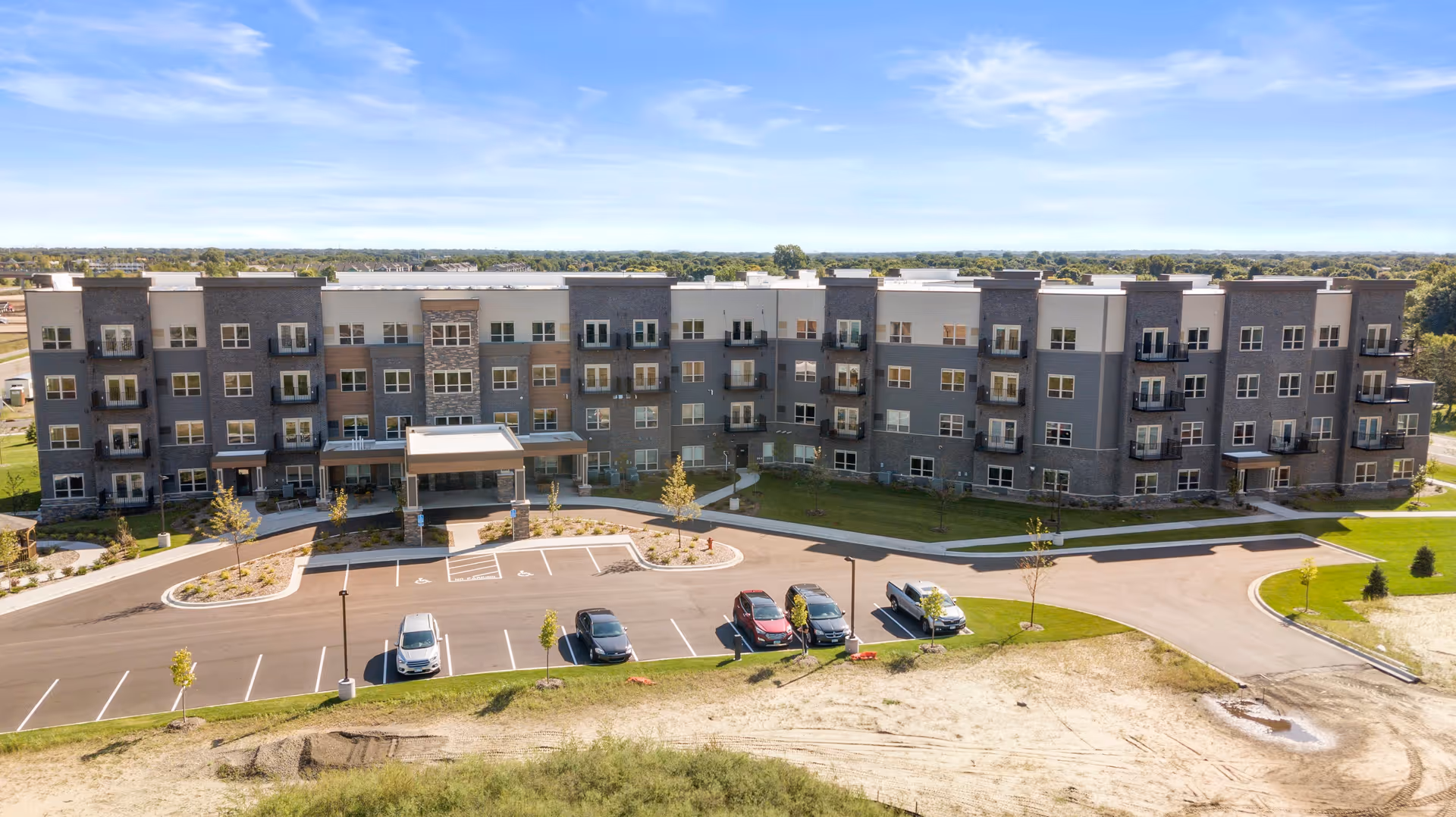 Exterior view of a modern, multi-story senior living facility building with multiple windows and balconies. There is a parking lot with several cars in front of the building, and a clear blue sky above.