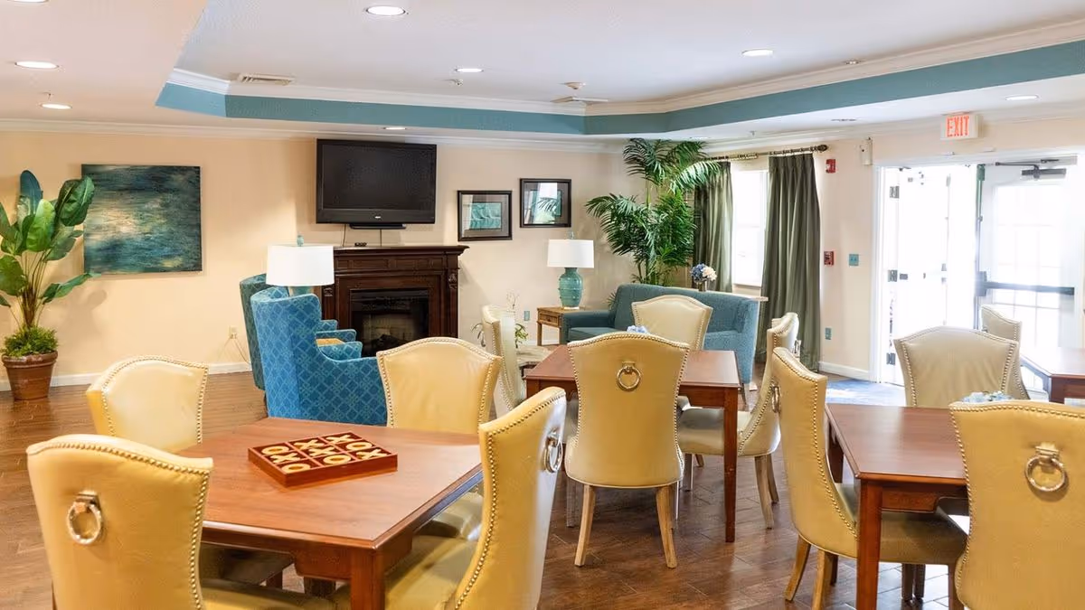 Bright communal dining and lounge area with multiple wooden tables and upholstered chairs, a TV over a fireplace, and potted plants.