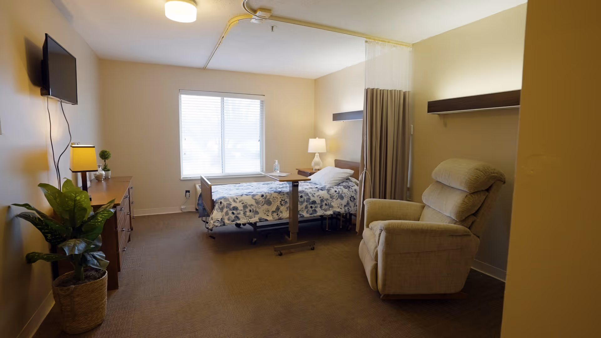 A well-lit senior living bedroom with a hospital-style bed, bedside table and lamp, recliner, dresser, wall-mounted TV and window.