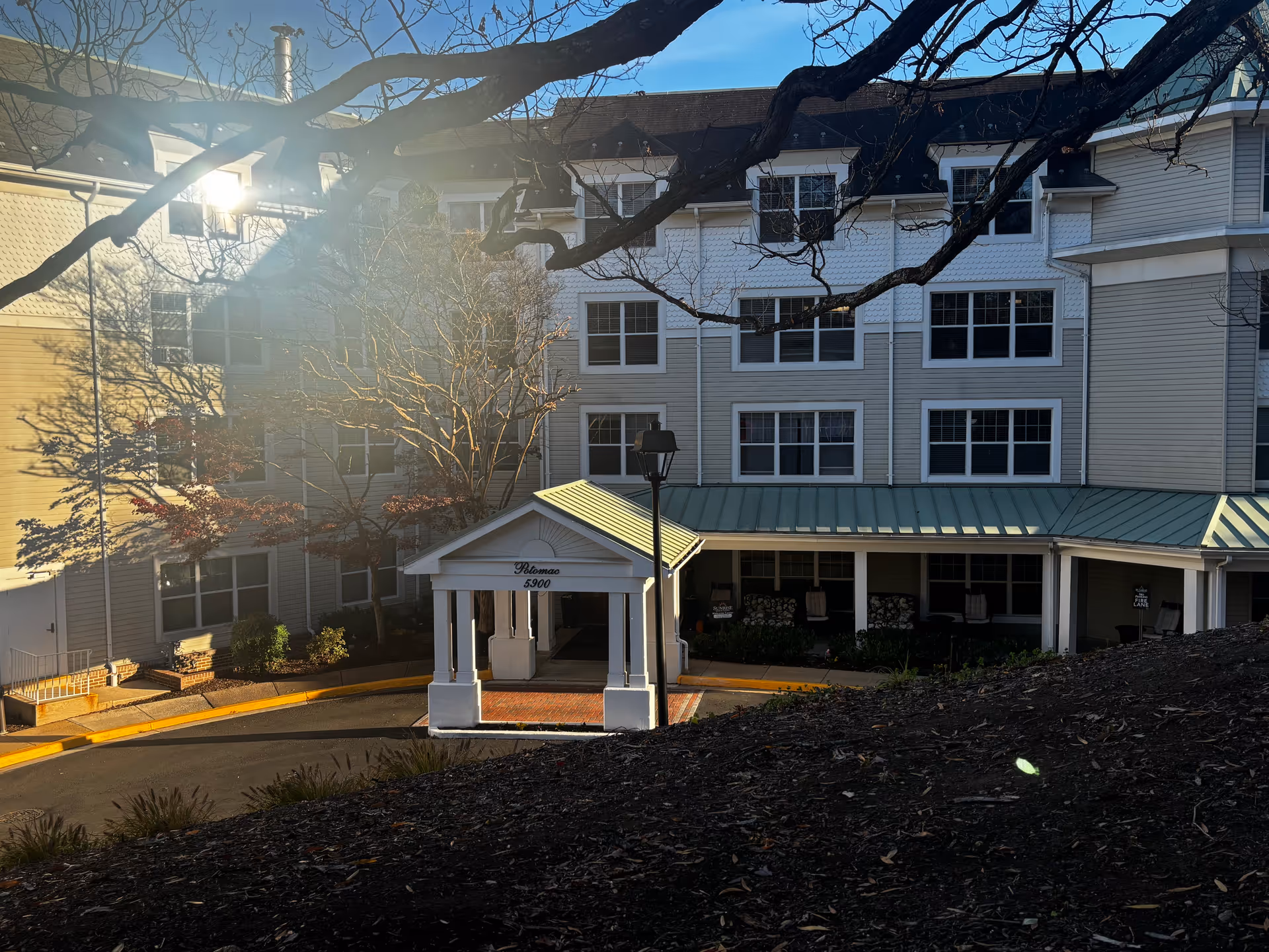 Exterior view of a senior living facility building with a covered entrance labeled 'Potomac 5900'. The building has multiple windows and a green metal roof over the entrance area. Bare tree branches are visible in the foreground with sunlight shining through.