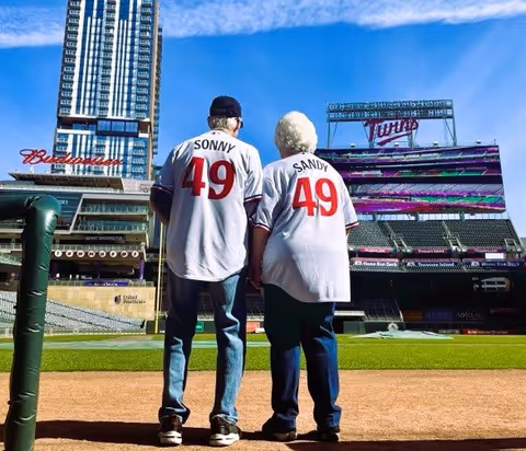 An elderly couple wearing matching white baseball jerseys with the names Sonny and Sandy and the number 49 stand side by side on a baseball field facing the stadium seating and scoreboard of the Minnesota Twins stadium on a sunny day.