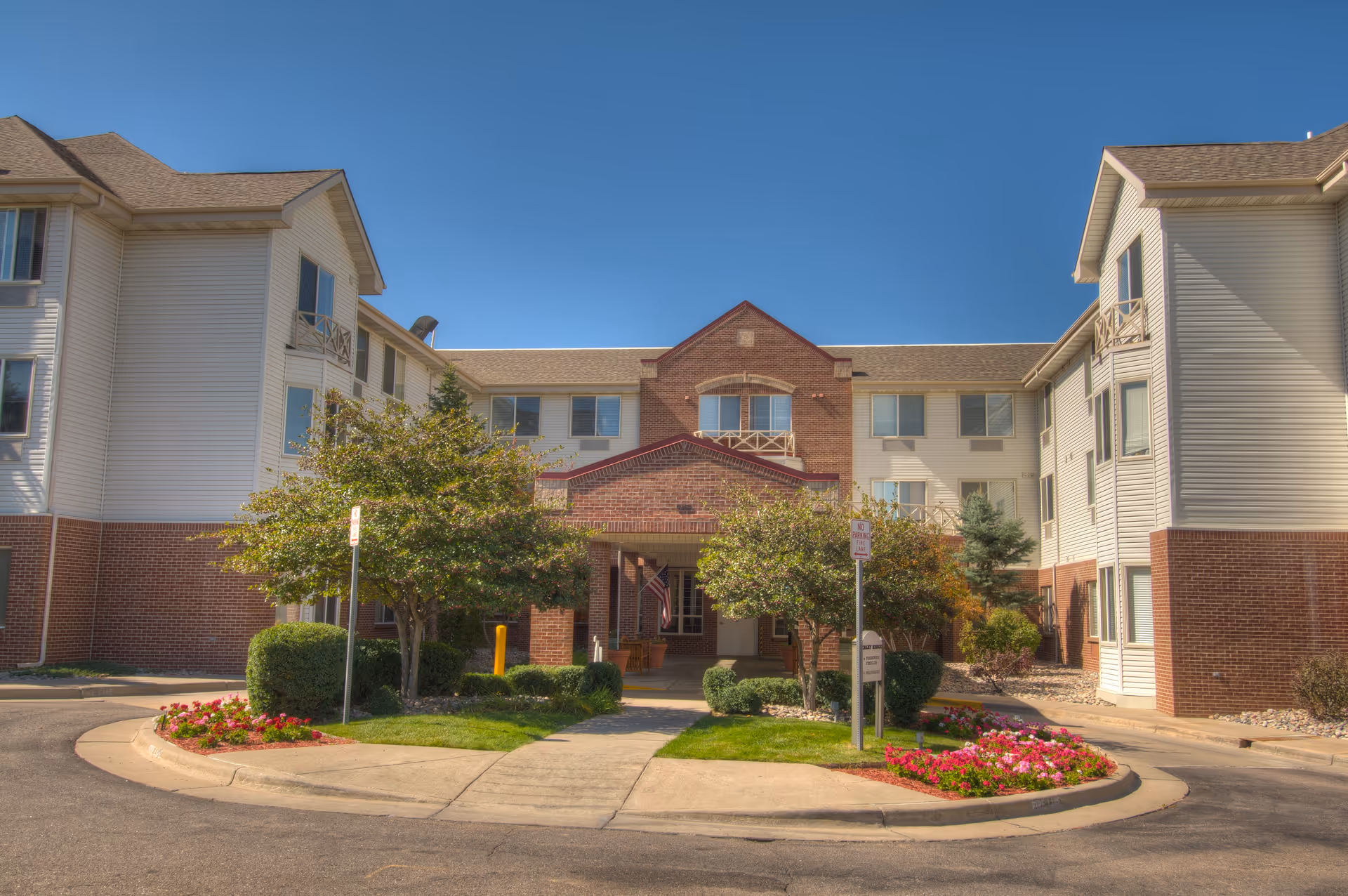 Front exterior view of Caley Ridge Assisted Living facility showing a three-story building with brick and light-colored siding, a covered entrance with an American flag, landscaped bushes, trees, and flower beds under a clear blue sky.