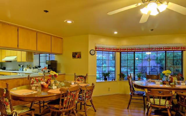 Bright communal dining area with wooden tables and chairs, a bay window with plants, and an open kitchen counter.