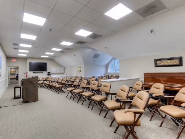 A large room with rows of beige cushioned chairs with attached writing surfaces arranged facing a podium and a large flat screen TV mounted on the wall. The room has a carpeted floor, white walls, a piano against one wall, and a window letting in natural light.
