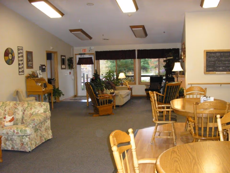 A cozy common area in an assisted living facility featuring wooden tables and chairs in the foreground, a floral upholstered armchair with a doll on it to the left, and a seating area with sofas and rocking chairs near large windows with dark valances. The room has soft lighting from ceiling fixtures and table lamps, with plants and framed pictures decorating the space.