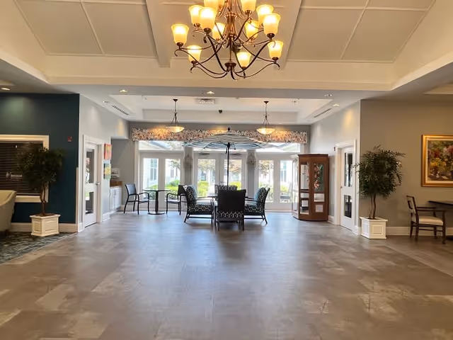 Spacious and well-lit common area in a senior living facility with a chandelier hanging from the ceiling, several chairs and a table arranged near large windows that let in natural light. There are potted plants on either side and a wooden display cabinet on the right side. The walls are painted in neutral tones with some artwork and decorative elements.