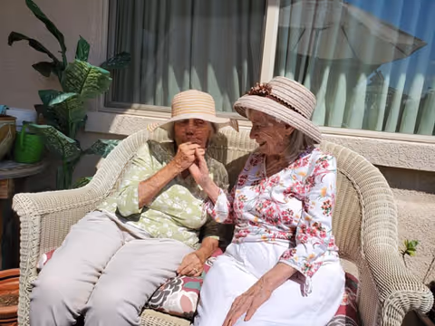 Two elderly women sitting closely together on a wicker bench outdoors, both wearing sun hats and light-colored clothing, with one woman holding the other's hand affectionately. There are plants and a window with curtains in the background.