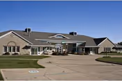 Exterior view of a single-story brick building with a gray roof under a clear blue sky. The building has multiple windows and a covered entrance with a driveway and landscaped grass areas in front.