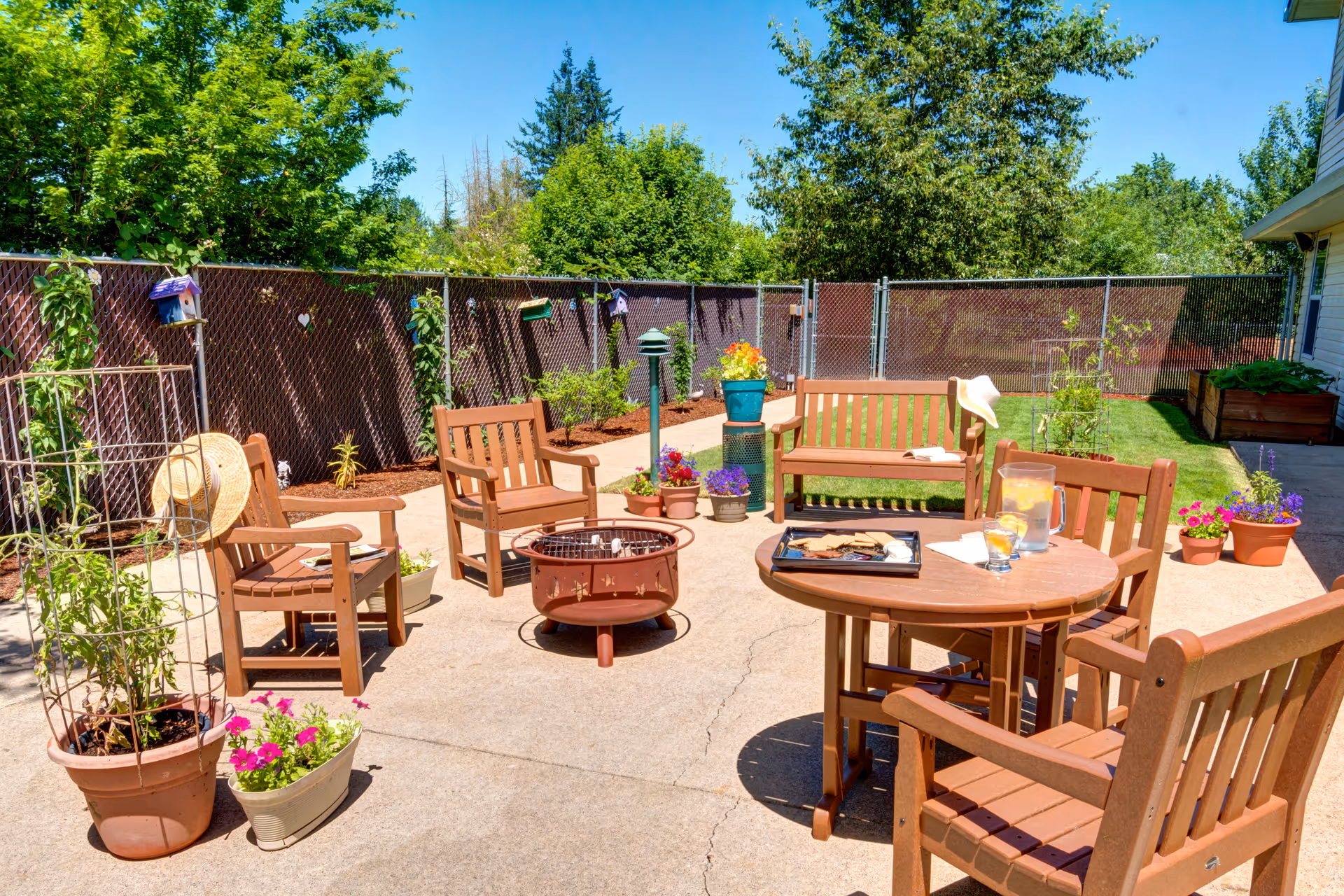 Outdoor patio area with wooden chairs and benches arranged around a fire pit and a round table. The table has a tray with snacks, a pitcher of lemonade, and glasses. Various potted plants and flowers are placed around the patio, with a chain-link fence and green trees in the background under a clear blue sky.