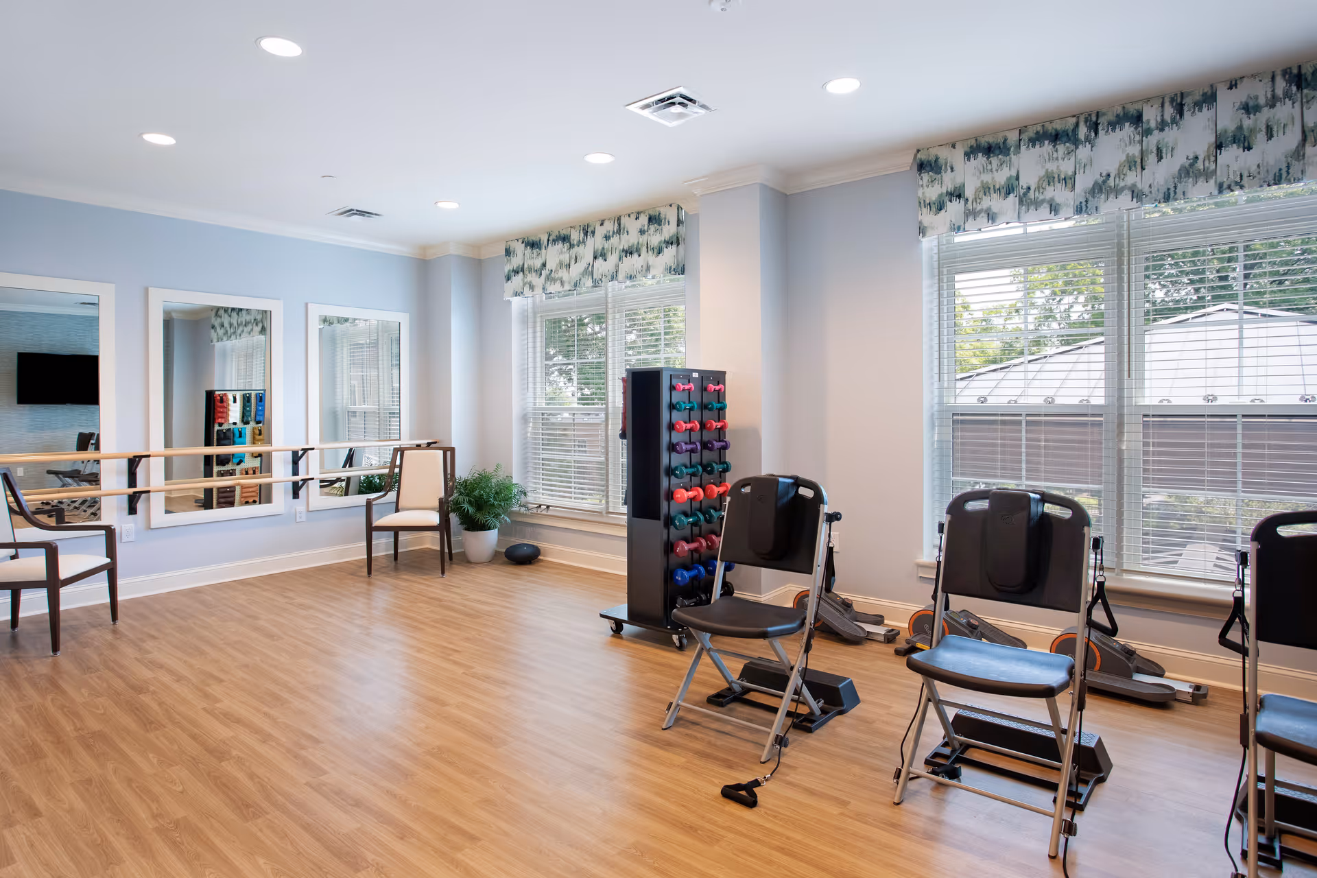 A bright exercise room with wooden flooring, light blue walls, and large windows with patterned valances. The room contains several black folding chairs equipped with resistance bands, a rack of colorful dumbbells, a ballet barre along one wall with three large mirrors above it, and a potted plant in the corner.