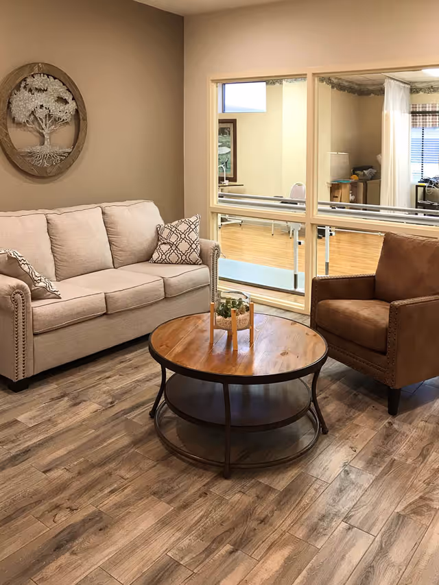 A cozy living room area with a beige sofa adorned with patterned pillows, a brown armchair, and a round wooden coffee table with a small decorative plant on it. The room has wood-look flooring and a wall decoration featuring a tree design. A large window reveals another room with wooden flooring and some furniture.