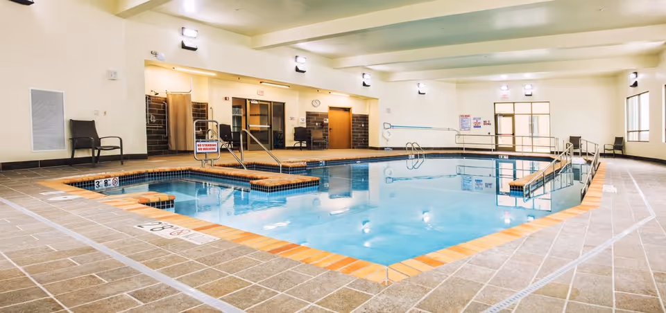 Indoor swimming pool area with clear blue water, tiled floor, handrails, and safety signs. The room has beige walls, multiple chairs along the walls, and several ceiling lights providing bright illumination.