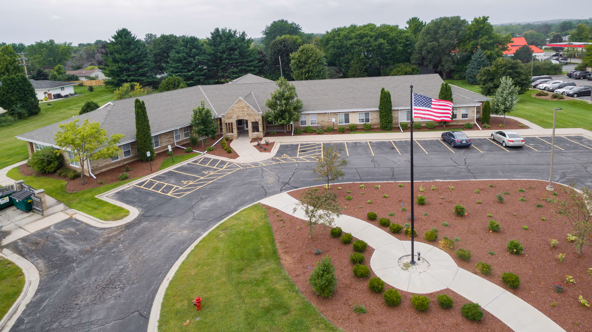 Aerial view of a single-story building with a gray roof and stone entrance, surrounded by landscaped greenery and a parking lot with several cars. An American flag is prominently displayed on a flagpole in a circular landscaped area in front of the building.