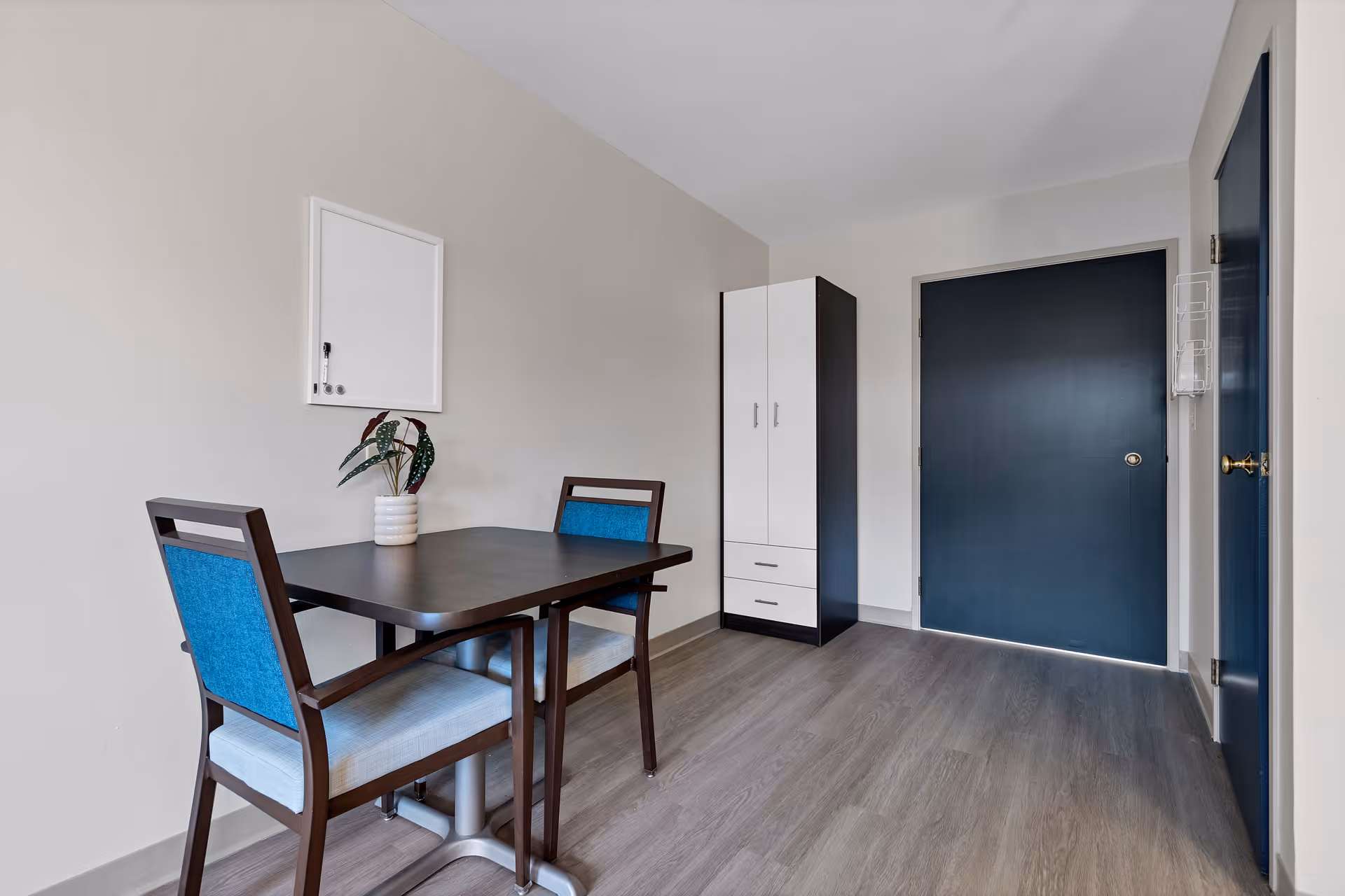 A small room with a dark wooden table and two chairs with blue cushions. There is a whiteboard on the wall above the table and a small potted plant on the table. In the background, there is a tall black and white cabinet and a dark blue door. The floor is covered with light wood laminate.