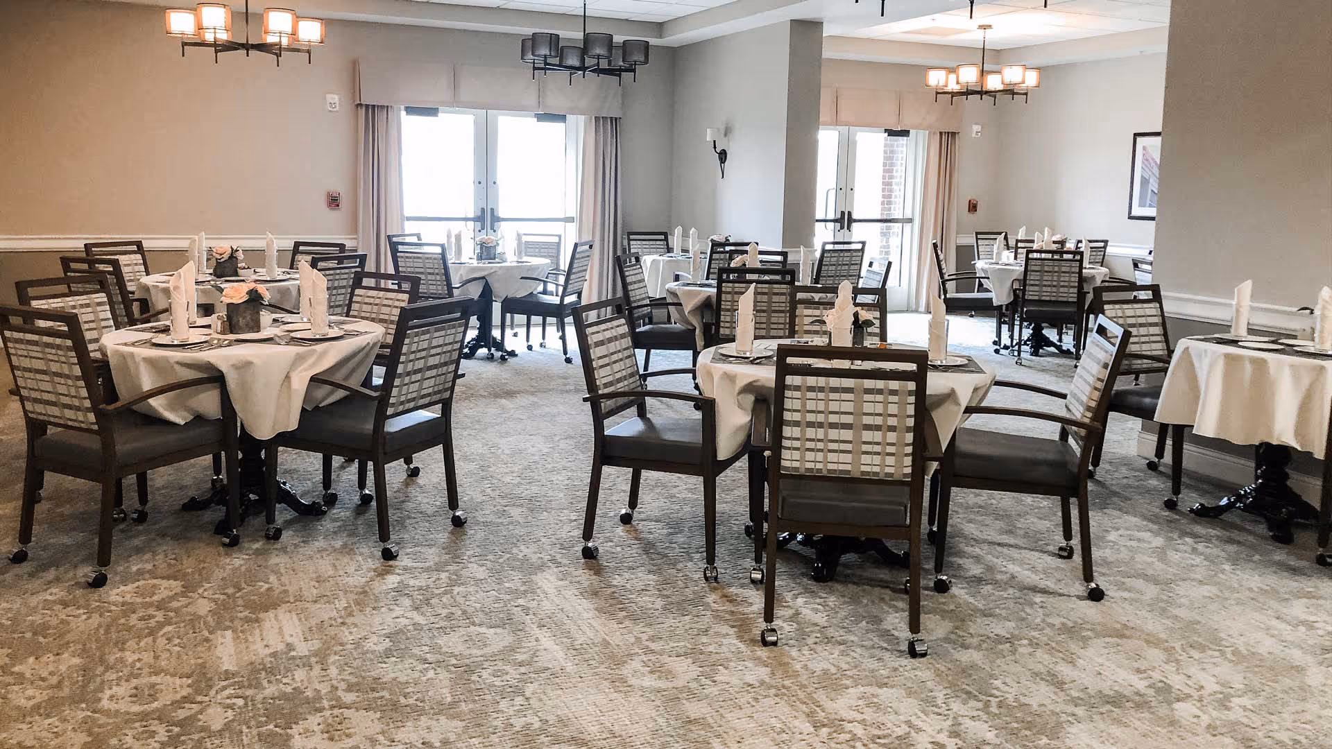 A dining room with multiple round tables covered with white tablecloths, each set with napkins and tableware. The room has carpeted floors, beige walls, large windows with curtains, and modern light fixtures hanging from the ceiling.
