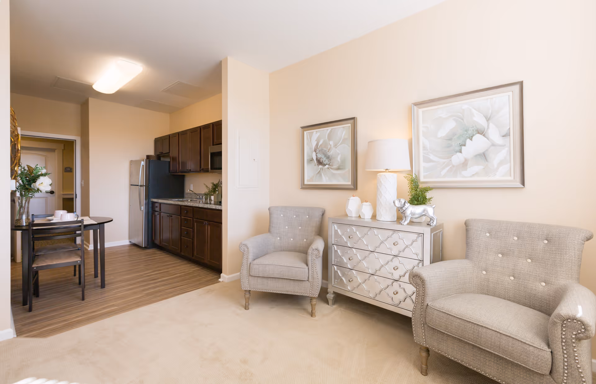 A cozy senior living interior featuring a small kitchen with dark wood cabinets and stainless steel appliances, a dining table with two chairs, and a sitting area with two beige upholstered armchairs, a mirrored chest with decorative items, and two floral paintings on the wall.