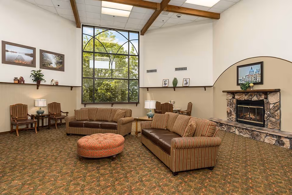 A cozy living room area with two striped sofas, a round patterned ottoman, two wooden chairs with cushions, two side tables with lamps, a large window showing green trees outside, and a stone fireplace with a framed picture above it. The room has a high ceiling with exposed wooden beams and patterned carpet flooring.