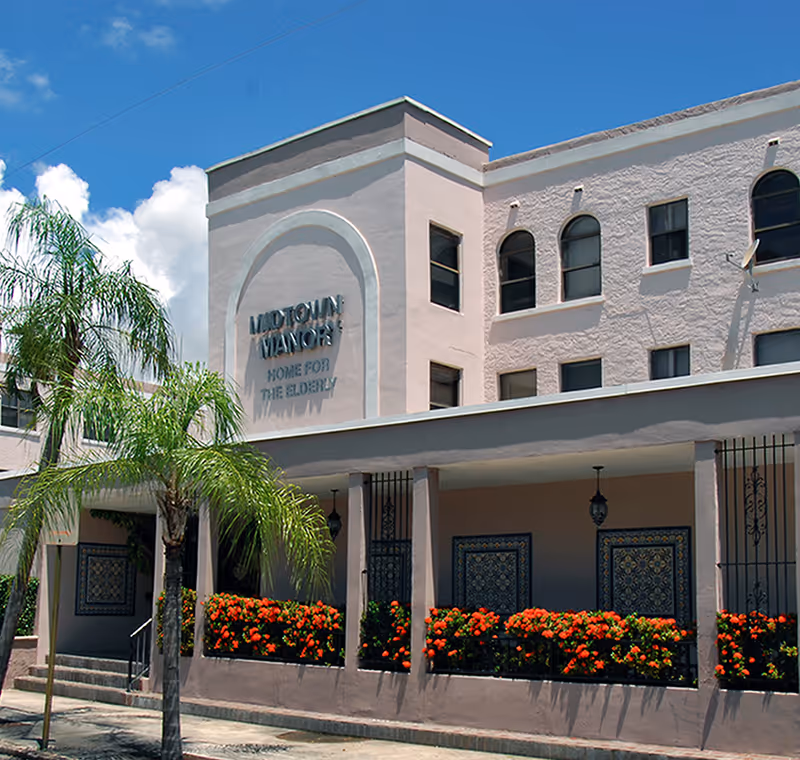 Exterior view of Midtown Manor, a senior living facility, showing a light pink building with multiple windows, decorative tiles, and a covered entrance with columns. There are palm trees and vibrant orange flowers in front of the building under a clear blue sky.