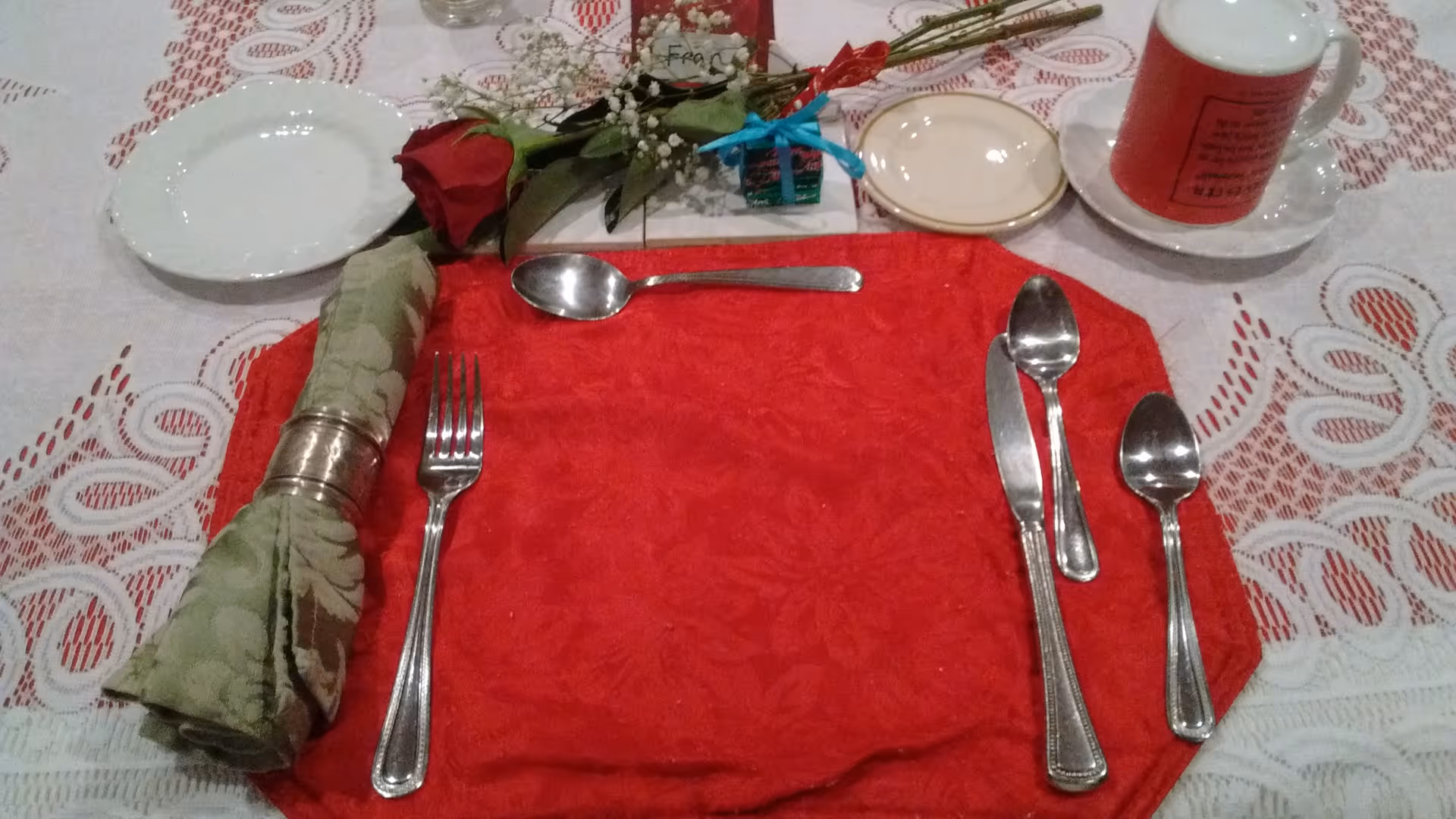 Close-up of a decorated table place setting with a red placemat, silverware, cloth napkin in a napkin ring, a rose, and a cup and saucer.