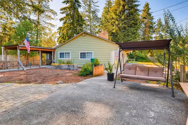 Outdoor view of a single-story yellow house with a covered porch displaying an American flag. The yard has a paved area with a cushioned swing bench under a canopy, some plants in pots, and tall trees in the background.