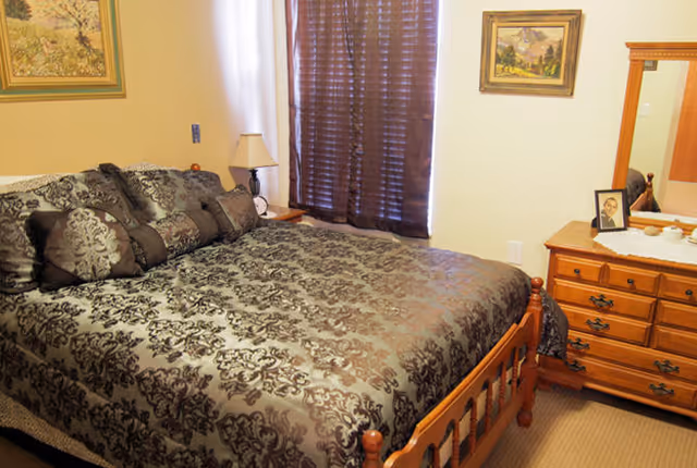 A bedroom with a wooden bed frame and a bed covered in a dark patterned comforter and multiple pillows. There is a wooden dresser with a mirror on the right side, holding a framed photo and some small items. A window with dark blinds is behind the bed, and two framed paintings hang on the walls.