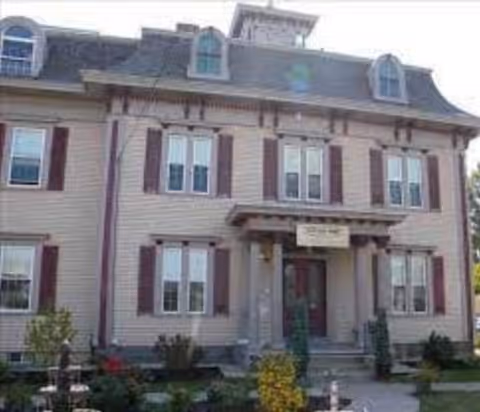 Front exterior view of a large, two-story house with beige siding and maroon shutters. The house has a central entrance with a small porch supported by columns, multiple windows on both floors, and a manicured garden with shrubs and flowers in front.