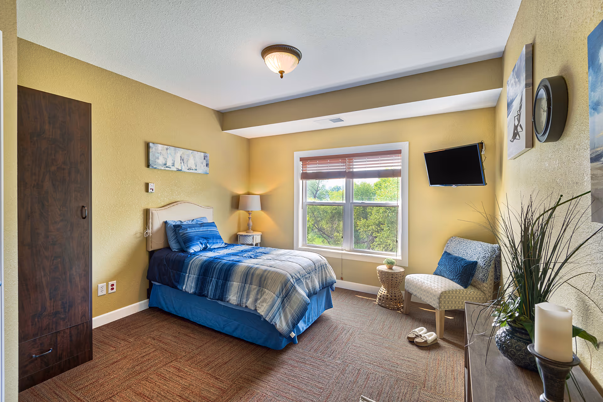 A cozy bedroom with a single bed dressed in blue and gray striped bedding, a wooden nightstand with a lamp, a window with blinds showing green trees outside, a wall-mounted TV, a cushioned chair with a blue pillow, a small round side table, slippers on the carpeted floor, and decorative plants and artwork on the walls.