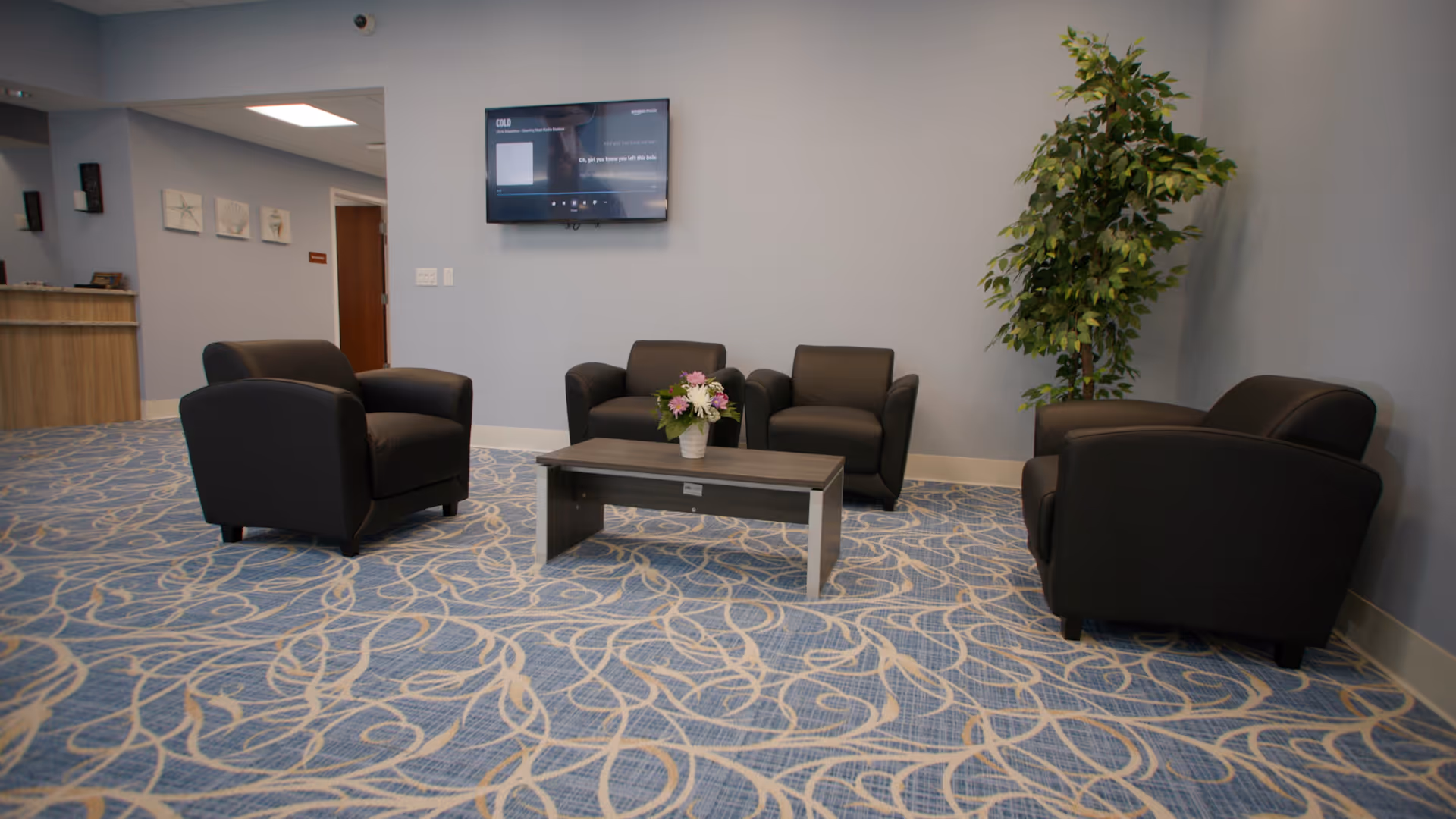 A seating area in a facility with four black armchairs arranged around a rectangular wooden coffee table with a flower arrangement on it. The room has blue patterned carpet, light blue walls, a flat-screen TV mounted on the wall, and a tall green plant in the corner.