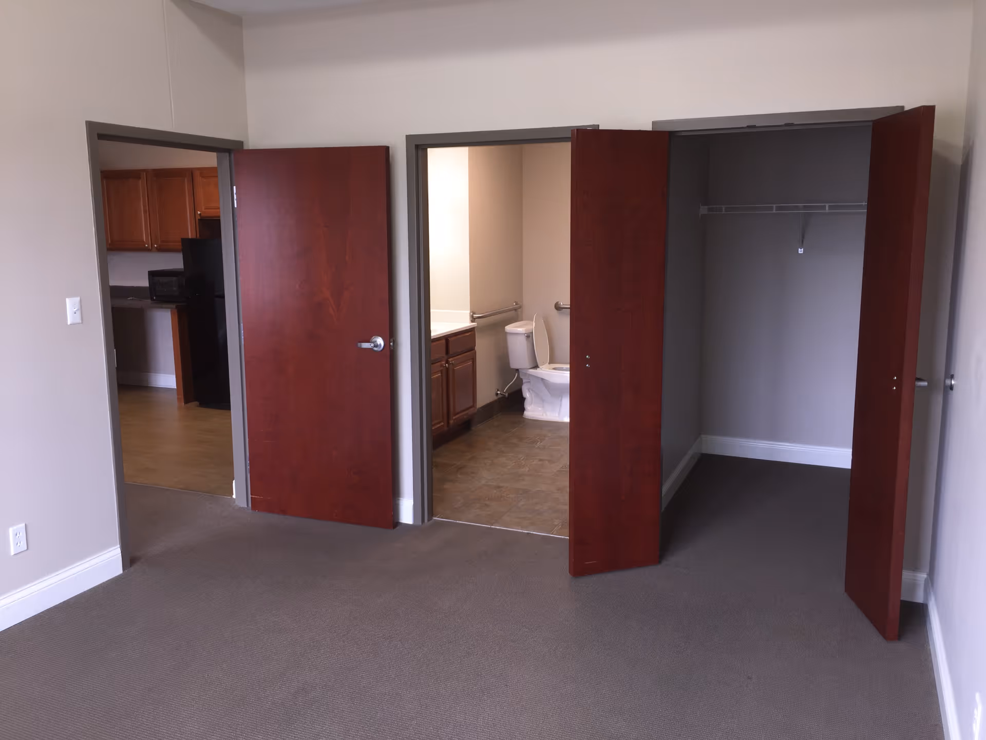 Carpeted apartment room with three open doors revealing a kitchen, a bathroom with a toilet and sink, and an empty closet.