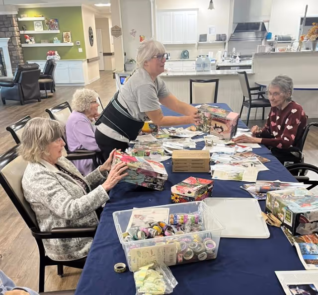 Seniors and a caregiver gathered around a table in a facility dining area working on craft projects with boxes and tape.