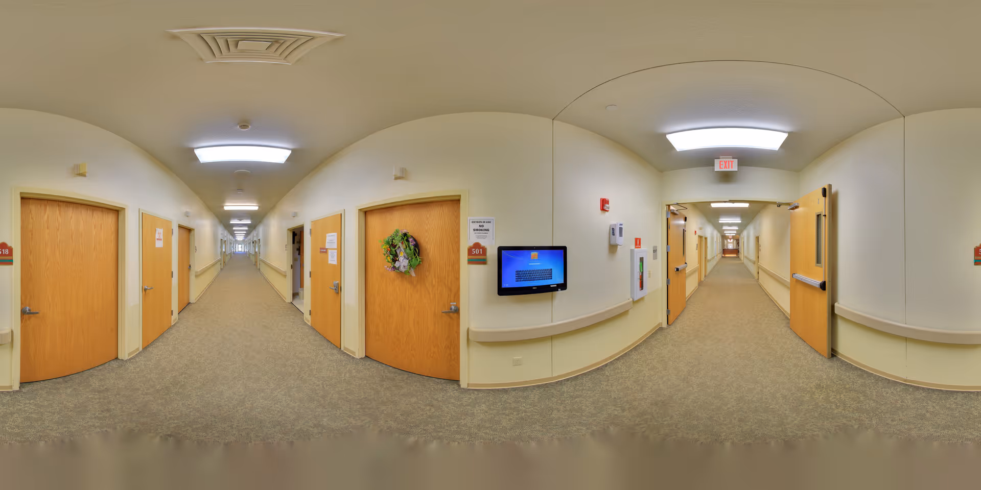 A long, well-lit hallway in a senior living facility with beige walls and carpeted floors. Multiple wooden doors line both sides of the corridor, some with room numbers and one decorated with a wreath. Handrails run along the walls, and ceiling lights illuminate the space. A wall-mounted screen is visible on the right side near an exit door.