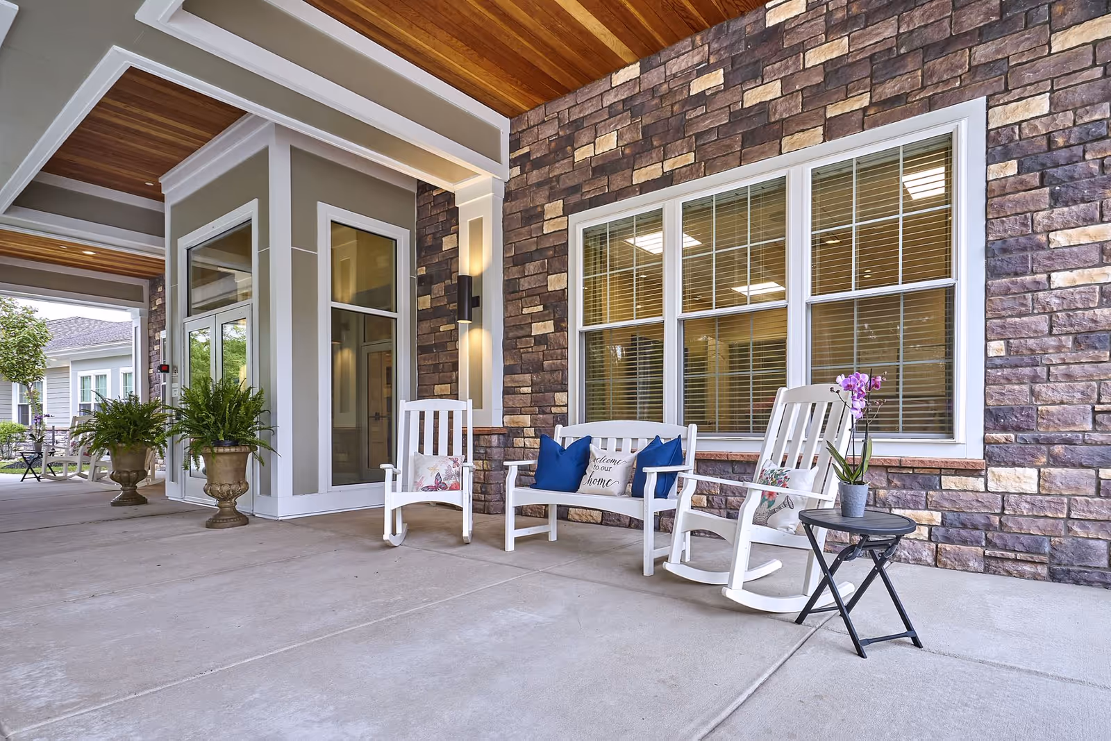 Covered outdoor seating area at a senior living facility with white rocking chairs and a bench adorned with decorative pillows. A small black side table holds a potted orchid. The wall behind is made of stone bricks with large windows, and there are potted plants near the entrance.
