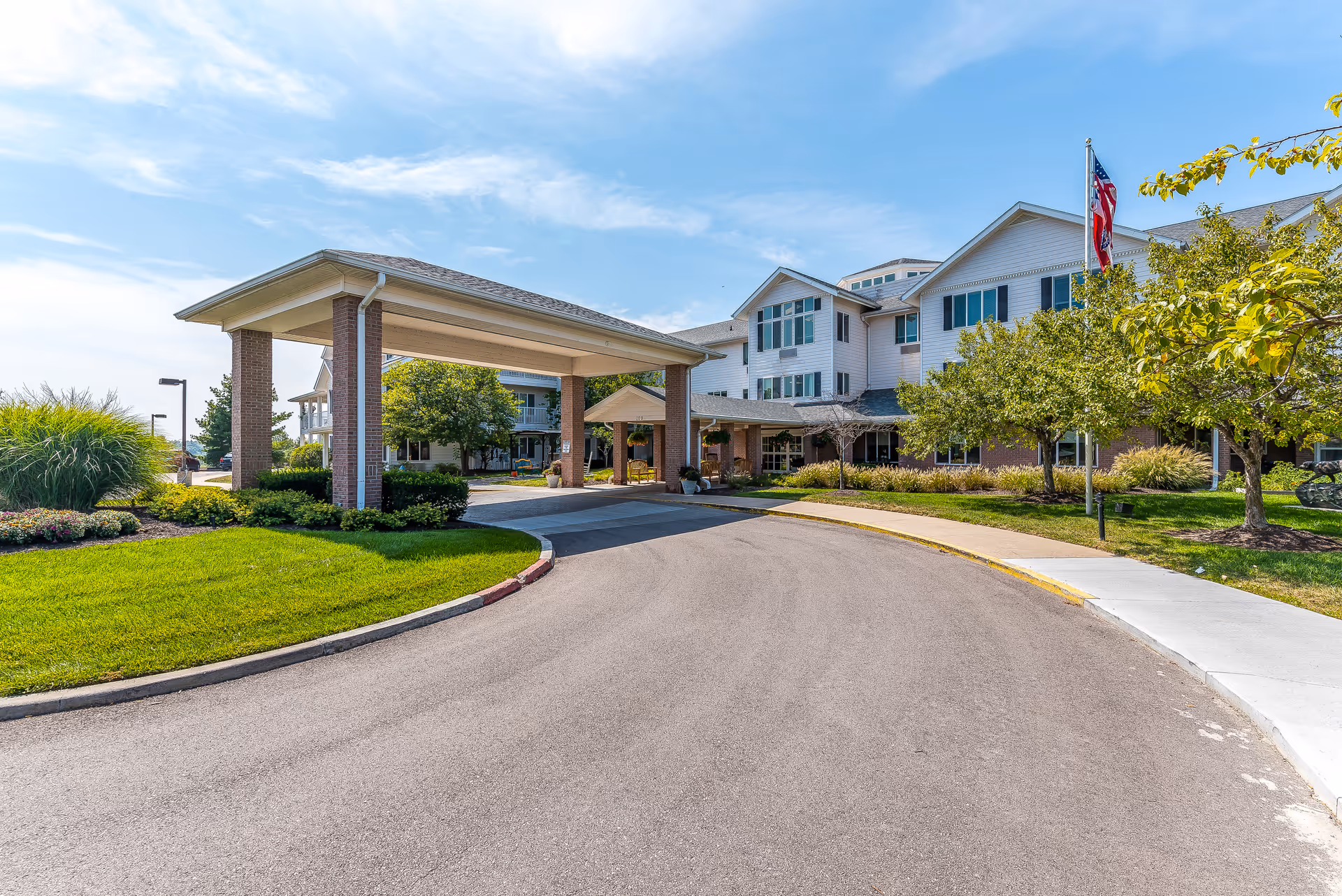 Exterior view of Solstice Senior Living at Columbia showing the main entrance with a covered driveway, a curved road, well-maintained green lawns, trees, and a clear blue sky.