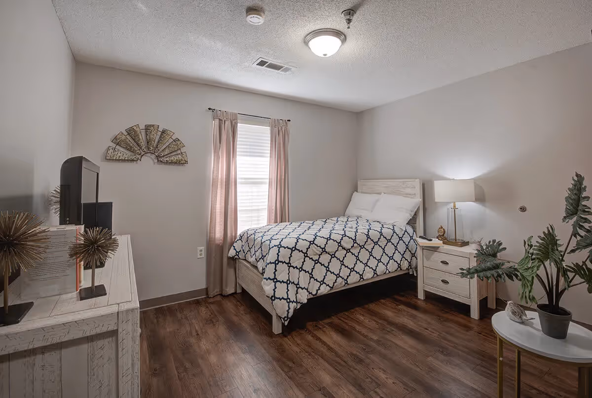 A cozy bedroom with a bed covered in a white and navy patterned comforter, two white pillows, a wooden nightstand with a lamp and clock, a window with light pink curtains, a wooden dresser with decorative items and a TV, a small round table with a potted plant, and wooden flooring.