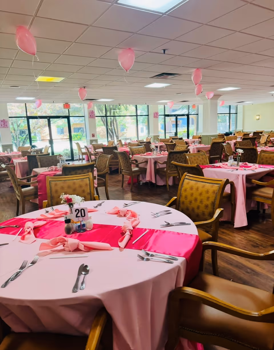 Dining room set for an event with round tables covered in pink tablecloths, chairs, and pink balloons.