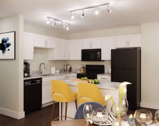 Modern kitchen with white cabinets, black appliances including a refrigerator, stove, microwave, and dishwasher. A kitchen island with a marble countertop has two yellow bar stools. The dining table in the foreground is set with plates, glasses, and a flower centerpiece. Track lighting is installed on the ceiling, and a piece of abstract art is hung on the wall.