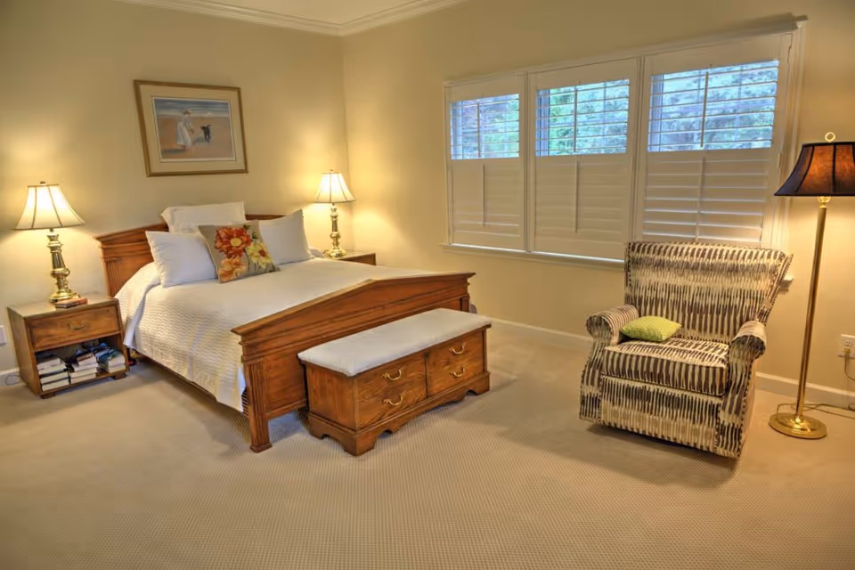 A cozy bedroom with a wooden bed frame, white bedding, and decorative pillows. There are two bedside tables with lamps on each side of the bed. A cushioned wooden bench with drawers is placed at the foot of the bed. A patterned armchair with a green pillow sits near a window with white plantation shutters. A floor lamp with a dark shade stands next to the armchair. The walls are painted light beige and a framed painting hangs above the bed.