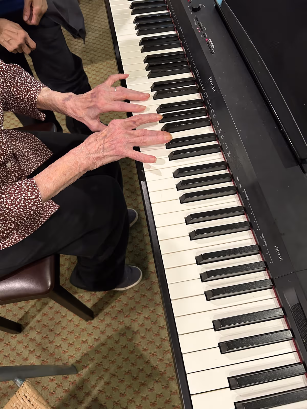 An elderly person playing a digital piano keyboard, with their hands positioned on the keys. The person is seated on a chair with a patterned carpet floor visible underneath.