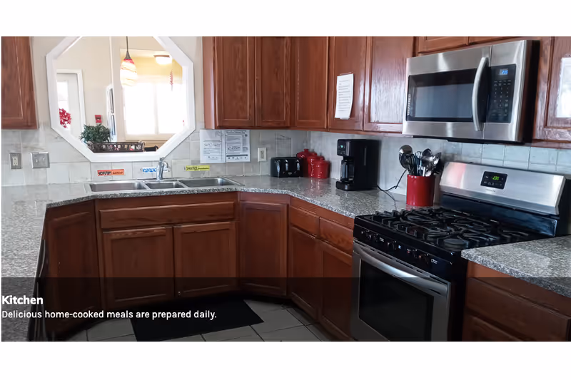 A kitchen with wooden cabinets, a granite countertop, a stainless steel microwave, a gas stove, a double sink, a coffee maker, a toaster, and kitchen utensils in a red holder. There is a window-like opening in the wall above the sink showing a bright room beyond.