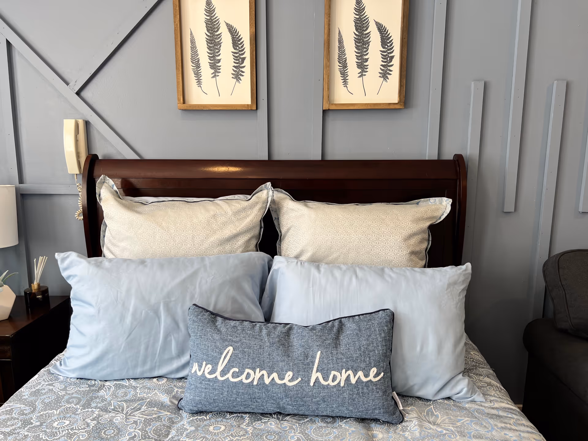 A neatly made bed with a dark wooden headboard against a gray paneled wall. The bed has multiple pillows including two large beige pillows, two light blue pillows, and a smaller blue pillow with the words 'welcome home' embroidered on it. Above the headboard are two framed botanical prints featuring fern leaves. A nightstand with a lamp and decorative items is partially visible on the left side.