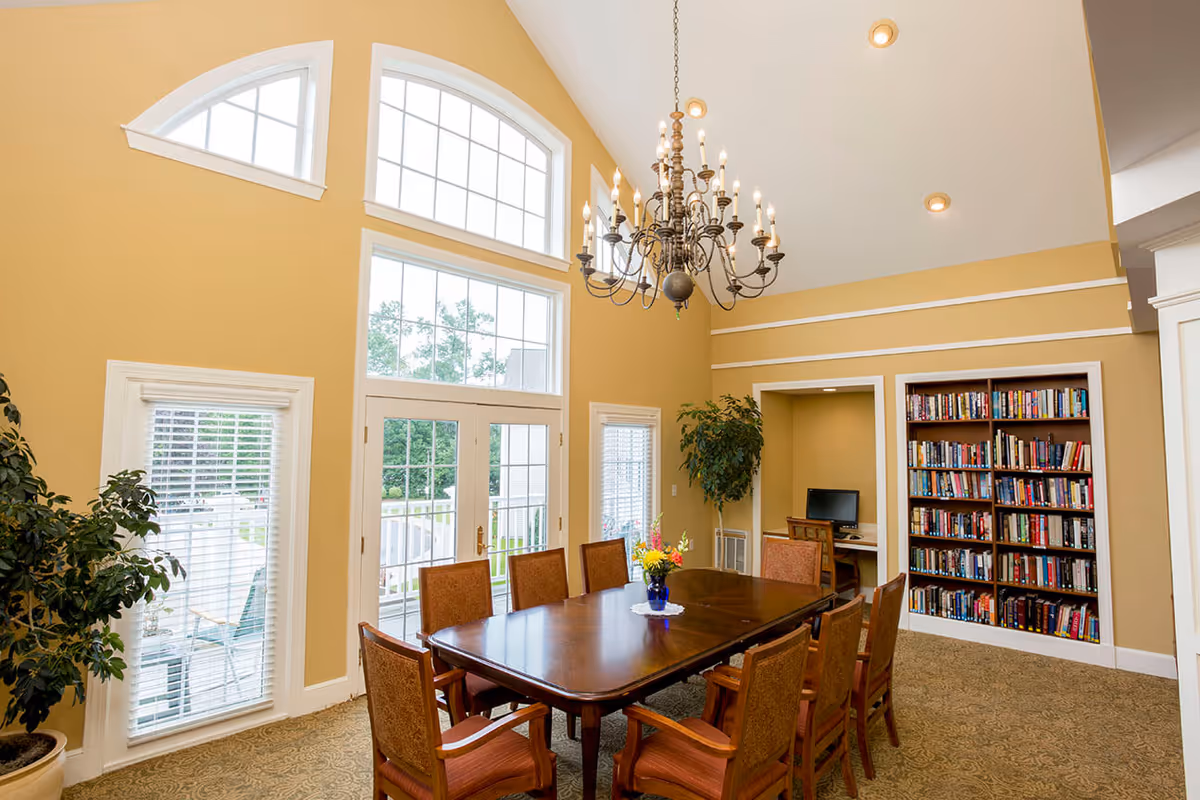 Well-lit communal dining room with a long wooden table and chairs, large arched windows and doors, a chandelier, and a bookshelf.