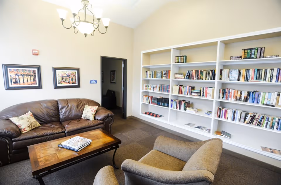 A cozy living room area with a brown leather sofa, two cushioned armchairs, a wooden coffee table with a book on it, and a large white bookshelf filled with books. The walls are light-colored with two framed pictures hanging above the sofa and a chandelier hanging from the ceiling.