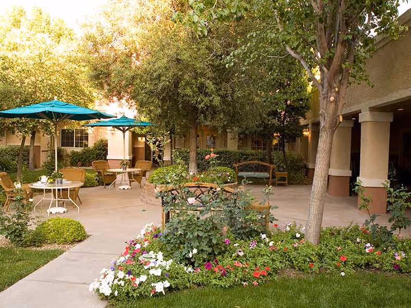 Outdoor patio area at Atria Seville with tables and chairs under teal umbrellas, surrounded by trees, flowering plants, and greenery. There is a paved walkway and a bench near the building entrance.