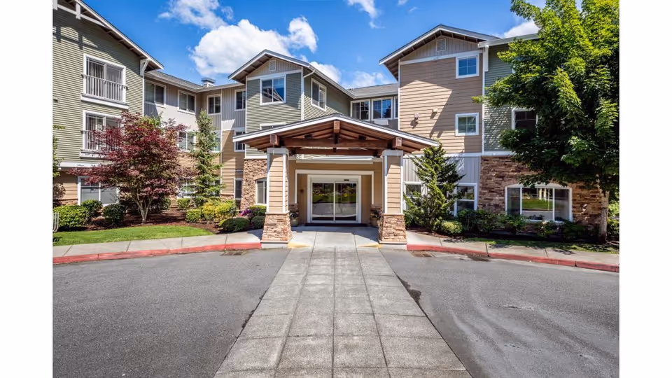 Front exterior view of a multi-story senior living facility named Woodland Terrace with a covered entrance, surrounded by trees and landscaping under a blue sky with some clouds.