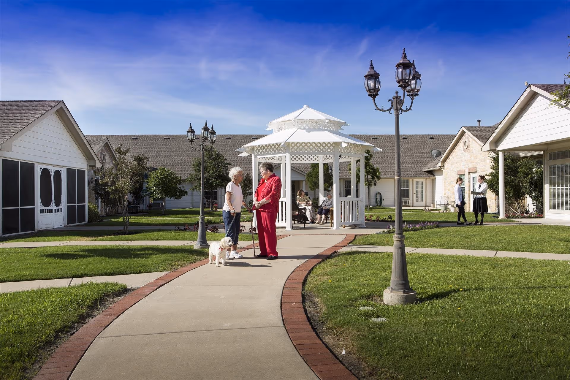 Two elderly women standing and talking on a paved pathway in a well-maintained garden area of an assisted living facility. One woman is holding a small white dog on a leash. In the background, there is a white gazebo with two people sitting inside and two other people standing and talking near the building. The sky is clear and blue.