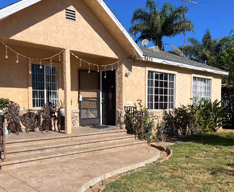 Exterior view of a single-story beige stucco building with a small porch featuring metal chairs and a table. There are steps leading up to the porch, windows with white frames, and some greenery including bushes and palm trees around the building under a clear blue sky.