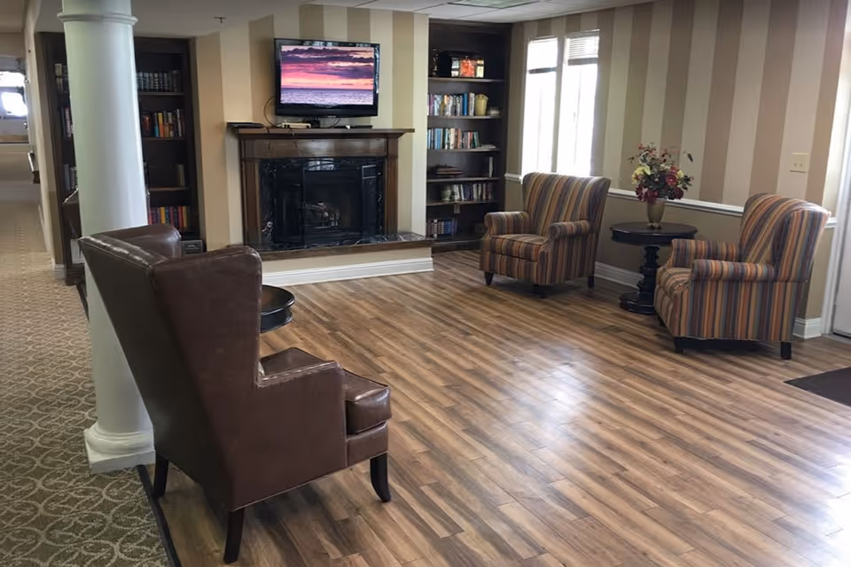 A cozy sitting area with two striped armchairs and one brown leather armchair arranged around a small round table with a vase of flowers. There is a fireplace with a TV mounted above it, flanked by built-in bookshelves filled with books. The room has wood flooring and beige striped walls, with a window letting in natural light.