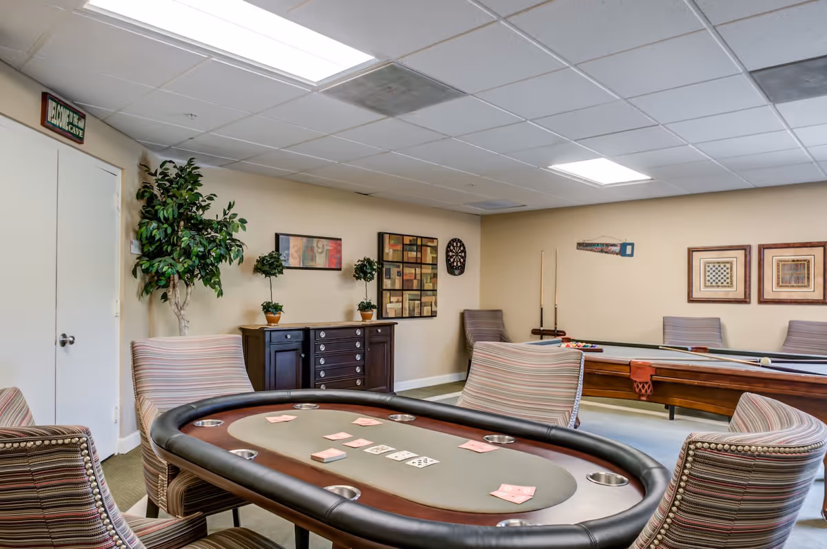 A recreational room with a poker table in the foreground surrounded by striped upholstered chairs. In the background, there is a pool table with cues on the wall and a dartboard. The walls are decorated with framed artwork and there is a potted plant near a door with a sign above it that reads 'Welcome to the Man Cave'.