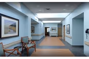 A wide hallway in a healthcare facility with light blue walls and ceiling tiles. The floor has a combination of wood and dark tiles. There are wooden chairs with cushions along the left wall, and framed landscape artwork hanging above them. The hallway is well-lit with recessed lighting and has handrails on both sides. At the far end, there is a doorway and additional framed pictures on the walls.